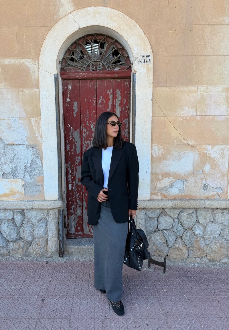 Blazer nero oversize, camicia bianca, gonna grigia e borsa nera con borchie. Lo sfondo presenta una porta di legno rossa e una parete di pietra texturizzata.