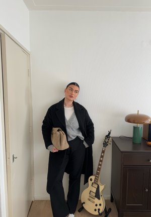 Abrigo negro largo, suéter gris, pantalones negros, sosteniendo una bolsa beige. De pie cerca de una lámpara marrón y una guitarra eléctrica en un soporte.