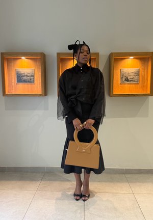 Mujer con vestido negro y top transparente sosteniendo un bolso marrón claro, entre dos cajas de madera para exhibición con fotos enmarcadas.