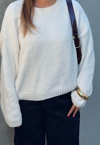 Άτομο που φοράει υφασμάτινο λευκό πουλόβερ, σκούρο παντελόνι, σκούρα τσάντα ώμου, χρυσό βραχιόλι και ροζ βερνίκι νυχιών στα δάχτυλα, δίπλα σε γκρίζο τοίχο.