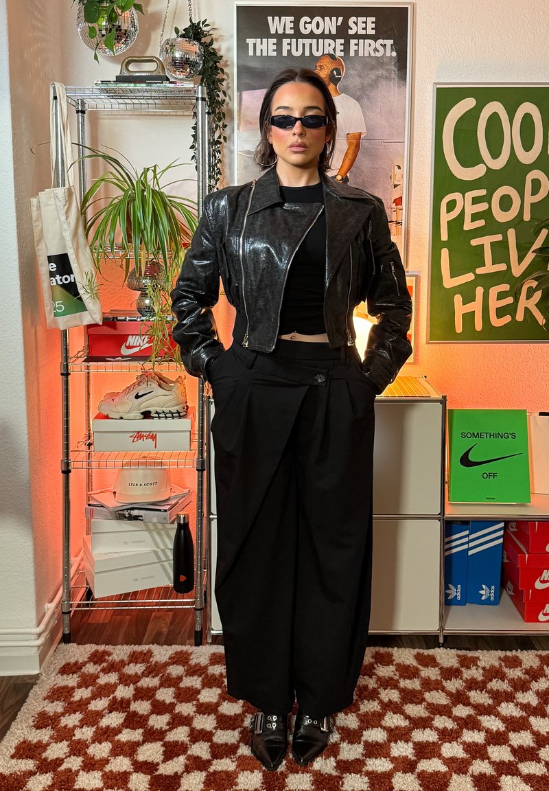 Veste en cuir noire brillante au design cropped, associée à un pantalon noir ample taille haute. Lunettes de soleil foncées et chaussures décoratives.