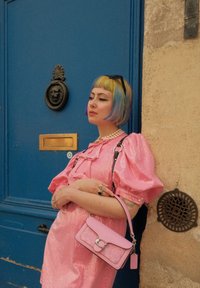 Ροζ φορεματάκι με λάμψη, φουσκωτά μανίκια και ραμμένες λεπτομέρειες; κρατά μία μικρή ροζ τσάντα με κούμπωμα; το φόντο περιλαμβάνει μία μπλε πόρτα και σιδερένιες λεπτομέρειες.