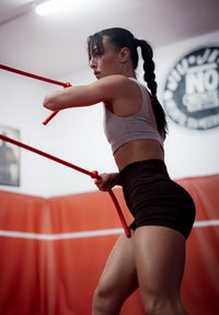 Športnik v svetlo sivi kratki majici in črnih kratkih hlačah, ki drži rdeče trening palice, na rdečem podloženem telovadnem ozadju.