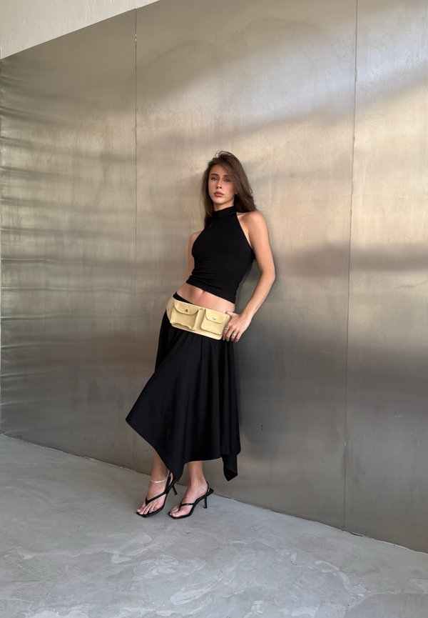 Femme portant un haut sans manches noir et une jupe avec un sac ceinture beige, debout contre un mur métallique lisse avec des talons noirs à brides.