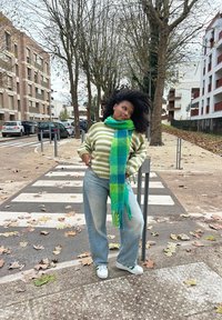 Ριγέ πουλόβερ σε μπεζ και πράσινο, σα scarf σε μπλε και πράσινους καρό, ανοιχτό μπλε τζιν, λευκά αθλητικά παπούτσια, στέκεται σε πεζοδρόμιο γεμάτο φύλλα.