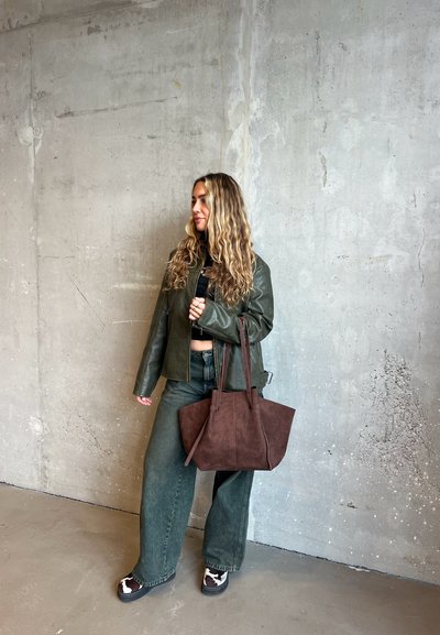 Chaqueta de cuero verde oscuro, blusa negra, jeans oscuros amplios y bolso de ante marrón. El calzado presenta una suela oscura y acentos de estampado de vaca claro.