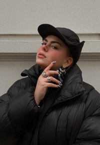 Casaco puffer preto com gola alta, boné preto combinando, joias ousadas incluindo anéis e grandes brincos de argola, e unhas com padrão.