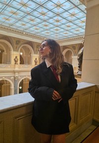 Czarna oversizeowa marynarka z długimi rękawami, biała koszula z guzikami i czerwona krawat. W tle widoczna ozdobna architektura i dekoracyjny sufit.