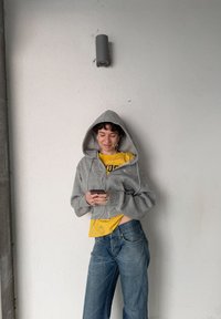 Sweat à capuche gris zippé avec une grande capuche, porté par-dessus un t-shirt jaune. Jean large bleu, personne tenant un smartphone, avec des écouteurs blancs visibles.