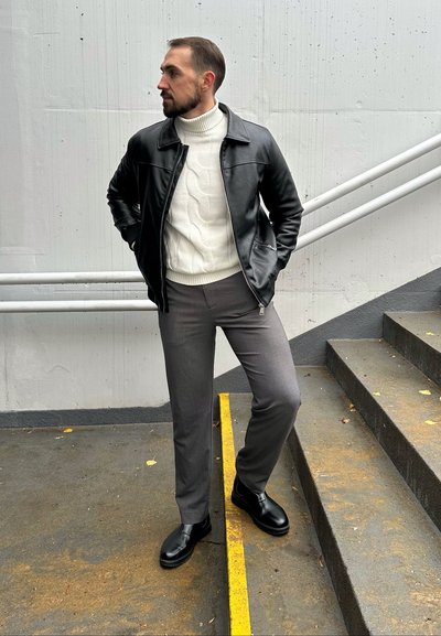 Chaqueta de cuero negra, suéter de cuello alto de punto trenzado blanco, pantalones de vestir grises y zapatos negros; de pie en unas escaleras con un fondo de hormigón.