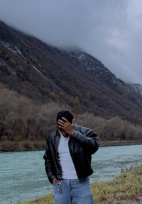Veste en cuir noire sur une chemise blanche, jeans bleus, debout près d'une rivière aux rives rocheuses, montagnes en arrière-plan, ciel nuageux.