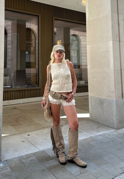 Top de tirantes de ganchillo beige, pantalones cortos de encaje blancos, botas altas de flecos beige. Una gorra beige y un bolso cruzado con un acento peludo completan el atuendo.