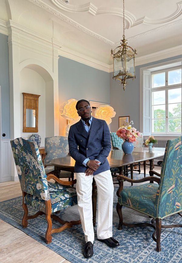 Hombre con blazer azul y pantalones blancos de pie junto a una mesa de comedor de madera con sillas florales en una habitación luminosa con ventana grande y techo ornamentado.