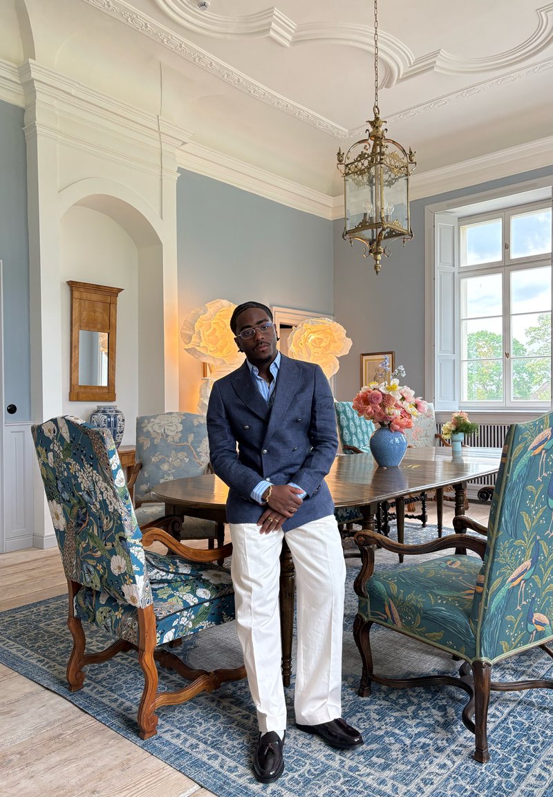 Homme en blazer bleu et pantalon blanc debout à côté d'une table à manger en bois avec des chaises à motifs floraux dans une pièce lumineuse avec une grande fenêtre et un plafond orné.