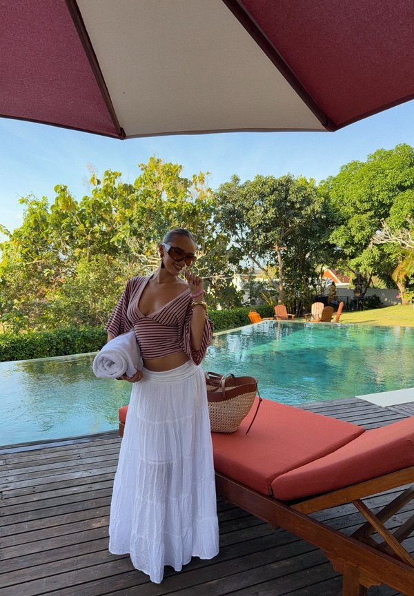 Mujer con gafas de sol sosteniendo una toalla junto a una piscina, de pie sobre una plataforma de madera cerca de una tumbona naranja bajo una gran sombrilla.