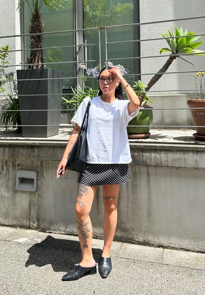 Bolso negro, camiseta de algodón blanca, falda corta negra con lunares, zapatos negros sin cordones. Tatuajes visibles en las piernas, de pie al aire libre cerca de plantas.