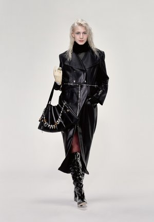 Black leather trench coat with a tailored fit, silver zipper, and belted waist. Paired with a handbag adorned with decorative accents and knee-high boots.