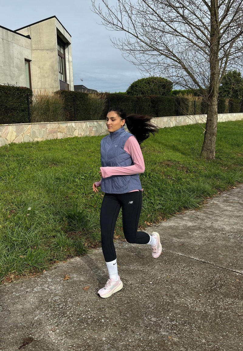 Corredora vestindo um colete cinza claro sem mangas por cima de uma camisa rosa de mangas compridas, leggings pretas e ténis rosas, a correr num caminho.