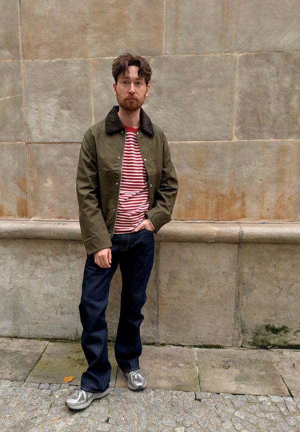 Hombre con cabello castaño y barba, de pie junto a una pared de piedra, vestido con chaqueta verde, camisa de rayas rojas y blancas, vaqueros oscuros y zapatillas grises.