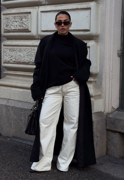 Abrigo negro largo, suéter negro ajustado y pantalones blancos holgados. Los accesorios incluyen gafas de sol oscuras y un pequeño bolso negro. Fondo texturizado.