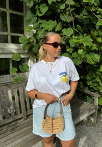 Witte grafische t-shirt met een gele citroenafdruk, lichtblauwe denimrok met rafelrand, geweven beige handtas en donkere zonnebril.