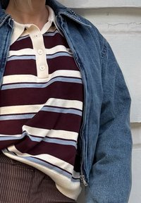 Henkilö, jolla on ylläsininen farkkutakki maroon, valkoisen ja vaaleansinisen raidallisen polo-paidan päällä, ja ruskeat suorat housut, seisoo valkoista seinää vasten.