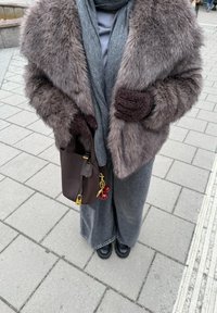 Brun fuskpälsjacka, grå scarf, grå topp, mörka vidbyxor, svarta stövlar, brun väska med gulddetaljer och körsbärsnyckelring fäst.