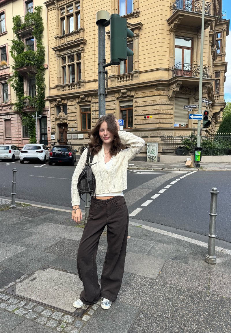 Cardigan blanc court en maille porté sur un haut simple, associé à un pantalon ample marron foncé et des baskets argentées. Sac noir et décor urbain en arrière-plan.