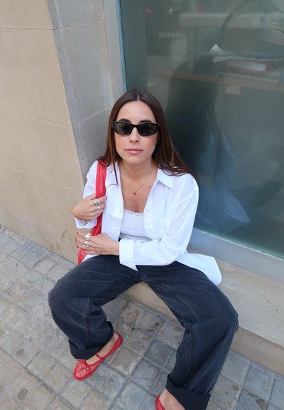 Camisa blanca de botones sobre una blusa de encaje, jeans negros de pierna ancha, unos zapatos planos rojos de trenzado y un bolso rojo; los accesorios incluyen anillos de plata apilados y gafas de sol.