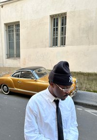 Beanie nero, occhiali trasparenti, camicia bianca e cravatta nera. Auto vintage dorata con tetto nero sullo sfondo contro un muro testurizzato.