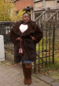 Femme portant un manteau en fourrure brun foncé, un haut blanc, une jupe en jean et des bottes marron, debout sur un trottoir près d'une clôture en fer forgé et d'un bâtiment en briques.