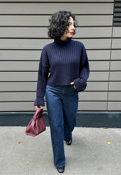 Mujer con pelo negro rizado, vistiendo un jersey de cuello alto de canalé azul marino, jeans de pierna ancha, zapatos negros, sosteniendo un bolso burdeos, caminando al aire libre.