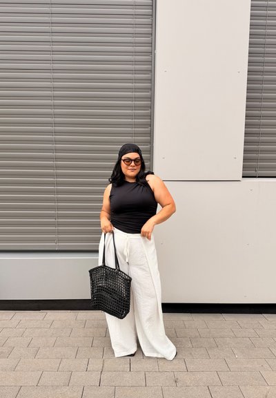 Mujer con top negro sin mangas, pantalones blancos de pierna ancha, pañuelo negro en la cabeza y gafas de sol sosteniendo un bolso tote negro tejido, de pie al aire libre.