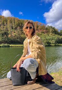 Saco de couro preto em forma de balde com uma alça curva, usado com jeans azul claro e uma jaqueta de corduroy bege, perto de um lago rodeado de árvores.