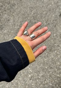 Zilveren ringen met gepolijste en gestructureerde afwerkingen zijn gedragen aan een hand, die een donker jack met een gele accenten manchet heeft.