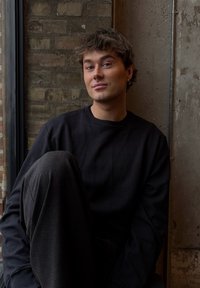 En person i en svart teksturert genser og mørke bukser sitter mot en murvegg, med en grå betongbakgrunn og naturlig lys fra et vindu.