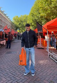 Afslappet outfit bestående af en sort zip-up jakke, lyseblå flared jeans og marineblå sneakers. Holder en orange tote bag med et logo.