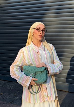 Mujer con pañuelo amarillo claro y camisa a rayas en tonos pastel sostiene un bolso acolchado verde con cadena plateada frente a una persiana metálica gris oscuro.
