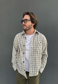 Chemise à carreaux boutonnée en beige et noir, en tissu tissé, associée à un t-shirt blanc, des lunettes de soleil foncées et un short vert olive.