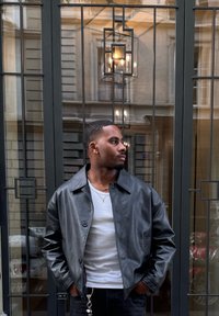 Mustast nahast jakk, millel on ees nööbid ja lai krae, kantud valge särgi üle, taustal tekstuuriga klaasaken geomeetriliste mustritega.