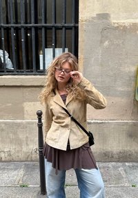 Blonde vrouw met krullend haar past haar bril aan en draagt een beige jas, een bruine top, blauwe jeans en een zwarte schoudertas op een stoep in de stad.