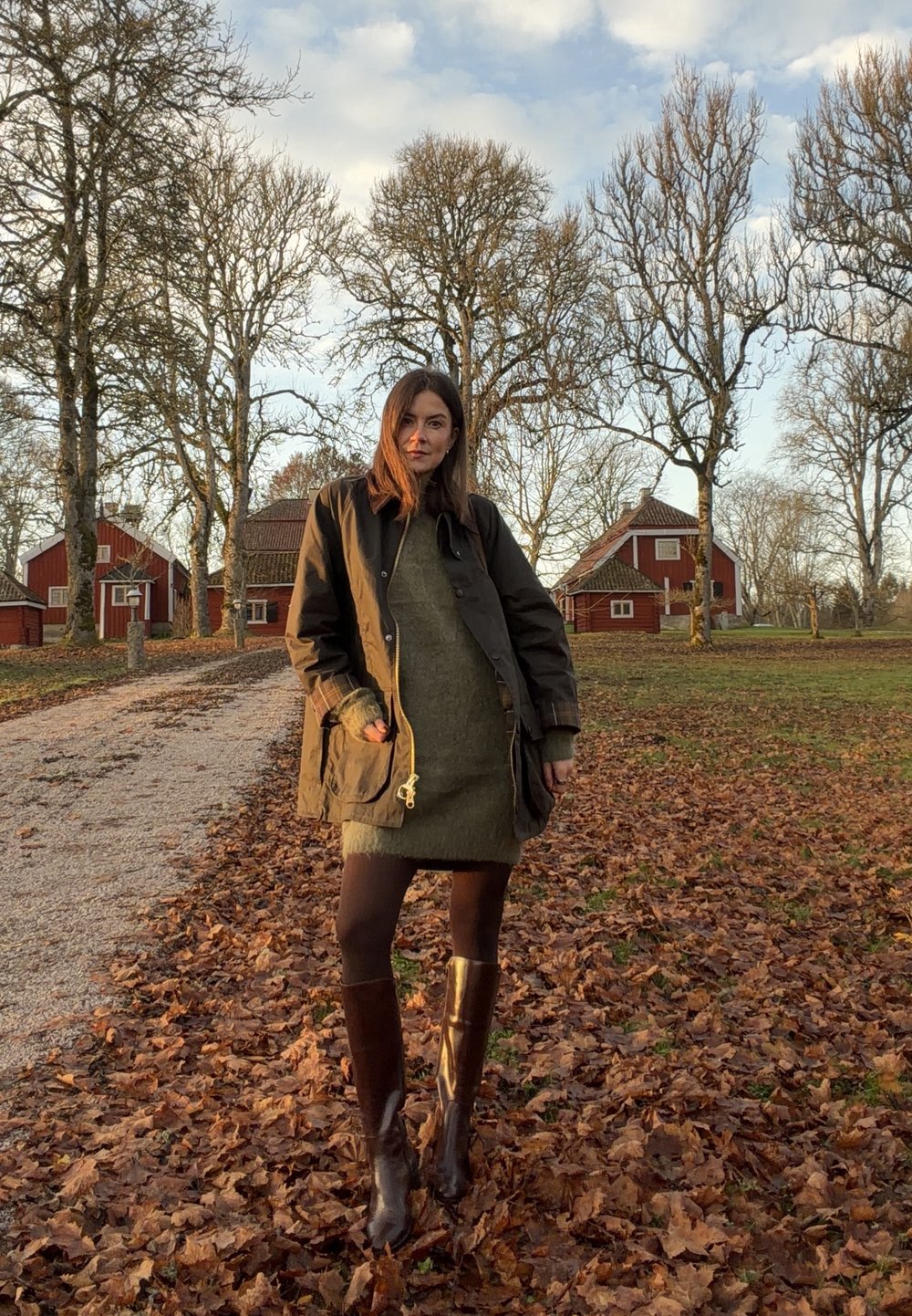 Khaki nepromokavý kabát se stahovací šňůrkou, tlusté olivově zelené šaty, hnědé punčocháče a hnědé kozačky po kolena, na venkovní cestě pokryté listím.