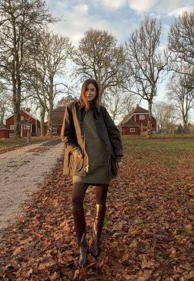 Abrigo impermeable color caqui con cordón, vestido grueso verde oliva, medias marrones y botas marrones hasta la rodilla, ambientado en un camino al aire libre cubierto de hojas.