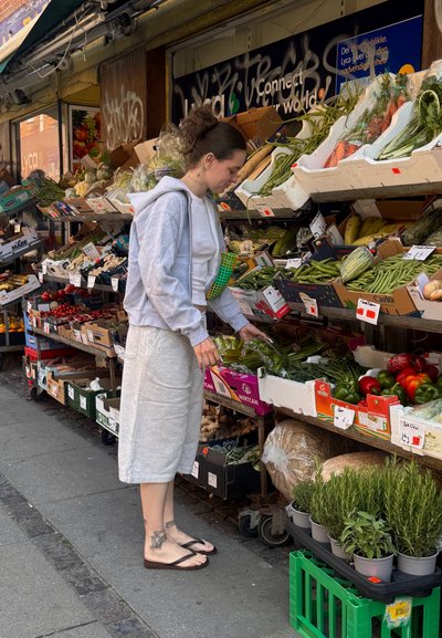 Persona examinando productos frescos en un mercado. Lleva un sudadera gris y pantalones cortos grises claros, de pie cerca de coloridas exhibiciones de verduras.
