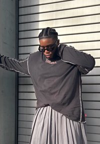 Čierna košeľa s dlhými rukávmi s bielym prešitím a malým logom, v kombinácii s plisovanou striebornou sukňou, na pozadí so sivými pruhmi.