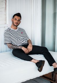 Schwarz-weiß gestreiftes T-Shirt mit einem kleinen roten Logo, schwarze Hose und schwarze Schuhe. Sitzt auf einer weißen gepolsterten Fläche.