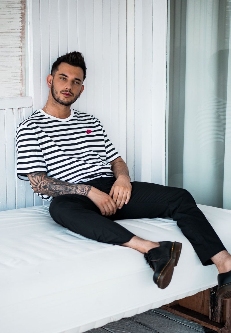 Schwarz-weiß gestreiftes T-Shirt mit einem kleinen roten Logo, schwarze Hose und schwarze Schuhe. Sitzt auf einer weißen gepolsterten Fläche.