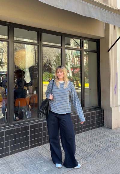 Mujer joven con cabello rubio, que lleva un suéter de rayas y pantalones de pierna ancha, está de pie en la acera frente a una cafetería con grandes ventanales y exterior revestido de azulejos.