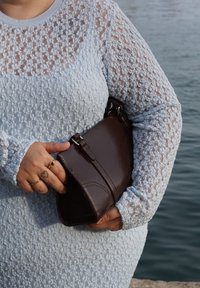 Mala de couro castanho com forma retangular e alça, segurada contra uma blusa de manga longa de tom azul claro com textura e padrão de renda.