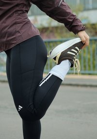 Leggings de sport noirs avec des rayures latérales, accompagnés de baskets marron et blanches, et d'une veste légère violette foncée.